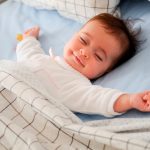 Baby smiling in bed with eyes closed and arms out.